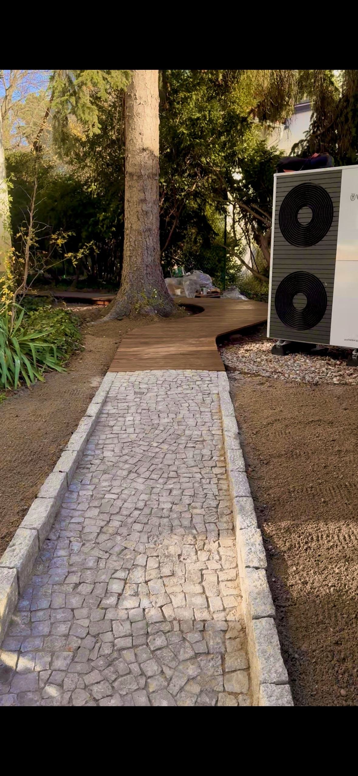 Geschwungener Holzsteg und Pflasterweg, harmonisch um den Baumbestand geführt.